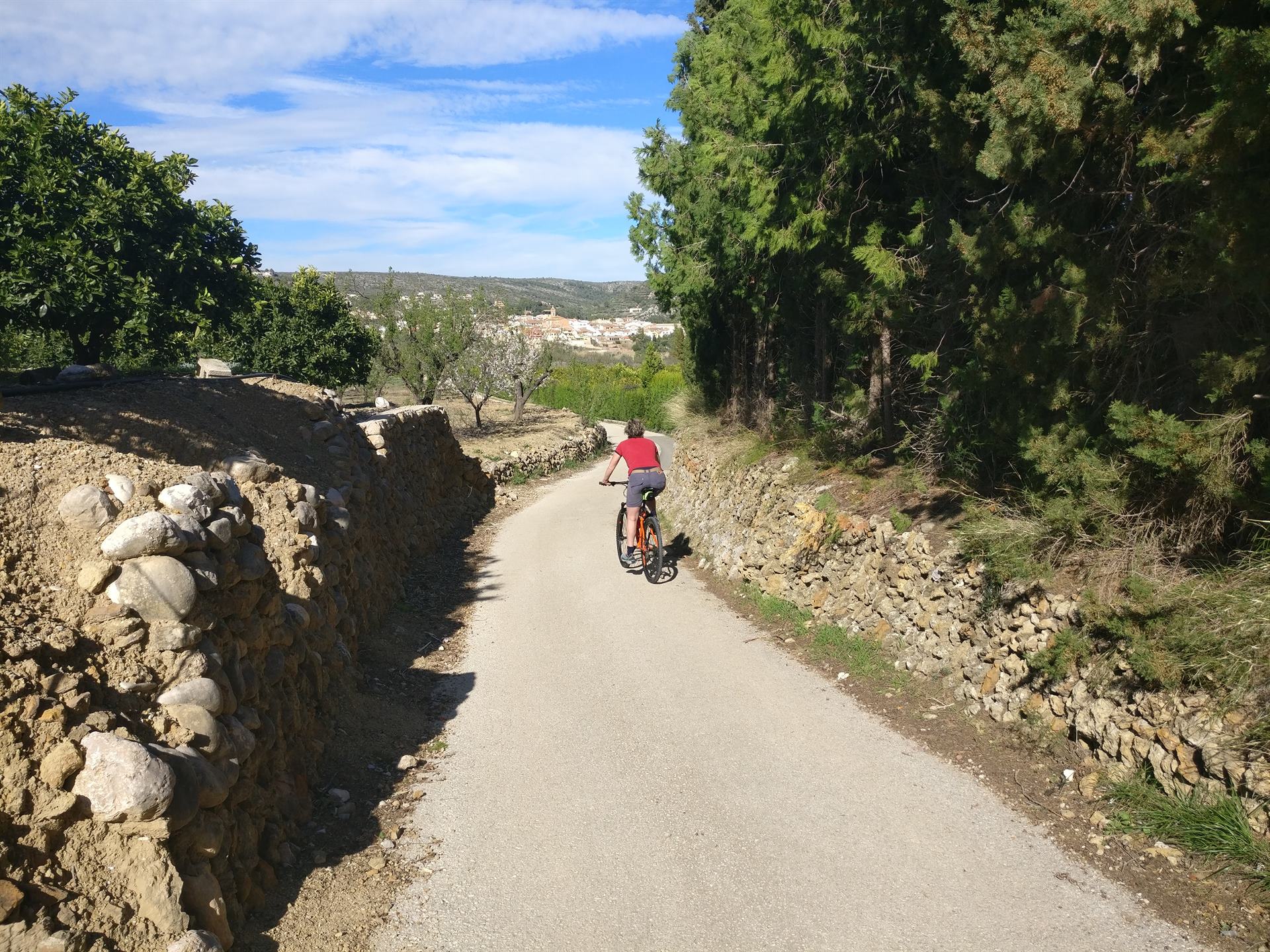 Mountain biker on a narrow concrete lane between dry stone walls, with the village of Alcalalí in the valley behind
